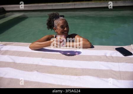 Femme adulte appuyée au bord de la piscine sur une serviette, tenant une boisson rose, lunettes de soleil sur la tête, smartphone à proximité Banque D'Images
