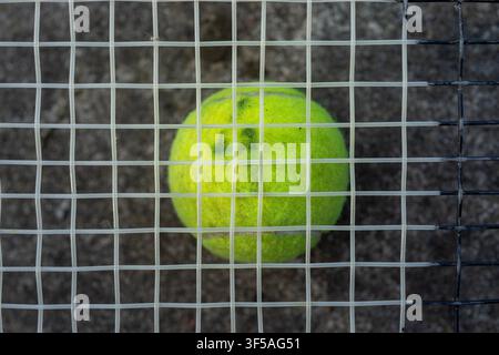 Balle de tennis vert néon posée derrière une grille de clôture métallique sous un éclairage extérieur, vue de près de face avec un fond de gravier flou et des barres d’acier verticales, créant un concept fort d’entraînement sportif et de barrière Banque D'Images