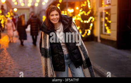 Femme souriante debout sur une rue pavée illuminée. Les lumières de Noël brillent derrière elle la nuit. Banque D'Images