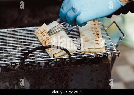 Main portant un gant bleu grillant des wraps de shawarma sur un grill métallique en plein air. La scène capture une cuisson active avec une chaleur visible et une atmosphère de street food décontractée Banque D'Images