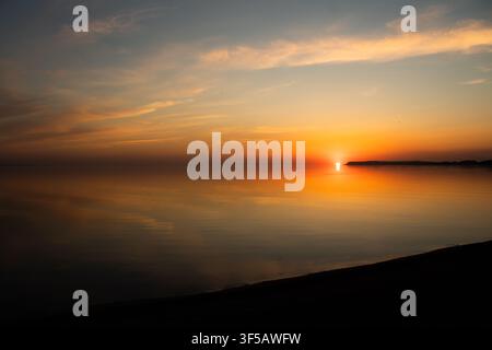 Soleil doré se couchant sur une eau calme avec un ciel orange se reflétant à l'horizon. Les silhouettes créent un paysage de coucher de soleil paisible et chaleureux Banque D'Images