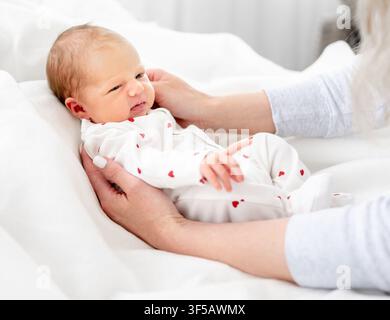 Des mains adultes tiennent doucement un nouveau-né vêtu d'une grenouillère blanche à imprimé de cœurs sur une parure de lit blanche et douce. Banque D'Images