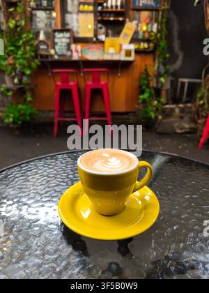 Un latte magnifiquement préparé servi dans une tasse jaune vif sur une table en verre, avec un art latte crémeux et un fond flou d'un café rustique et chaleureux Banque D'Images