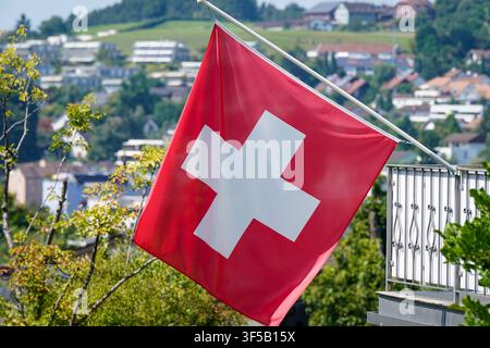 Drapeau national suisse flottant sur un mât devant un quartier résidentiel avec un paysage de collines en arrière-plan. Image symbolique de la Suisse, croix suisse, drapeau national, patriotisme, identité, État, nation, municipalité, zone résidentielle, architecture, paysage, été, vue extérieure, tourisme, Europe, panorama, lumière du jour. *** Le drapeau national suisse flotte sur un mât devant un quartier résidentiel avec un paysage de collines en arrière-plan. Image de stock : Suisse, croix suisse, drapeau national, patriotisme, identité, État, nation, municipalité, zone résidentielle, architecture, paysage, été, Banque D'Images