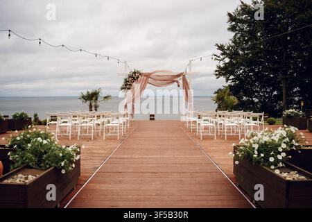 Installation pour une cérémonie de mariage sur la plage en plein air avec des chaises blanches et une arche fleurie surplombant l'océan. Lieu de mariage romantique sur la côte, mariage de destination tropicale, décoration élégante d'autel au bord de la mer Banque D'Images