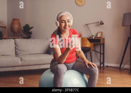 Femme âgée caucasienne heureuse écoutant de la musique tout en utilisant son smartphone à la maison, concept de perte de poids Banque D'Images
