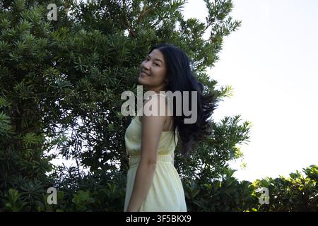 Femme adulte asiatique debout et souriant près d'un arbuste à feuilles persistantes dans le jardin, portant une robe jaune clair Banque D'Images