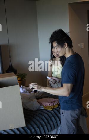Couple asiatique déballant des vêtements d'une grande boîte en carton sur un lit dans une chambre, tenant un smartphone Banque D'Images