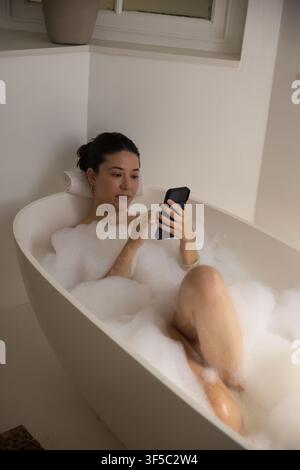 Femme allongée dans une baignoire indépendante avec de la mousse à bulles tenant un smartphone noir, tête sur une serviette roulée Banque D'Images