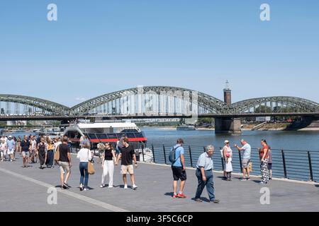 De nombreux piétons et touristes flânent par temps ensoleillé d'été sur la promenade riveraine du Rheingarten à Cologne. À l'arrière-plan, le célèbre pont Hohenzollern s'étend sur le Rhin, derrière lequel on aperçoit la tour de foire de Cologne-Deutz. À l'embarcadère se trouve un bateau de tourisme fluvial. Ce motif vivant symbolise le tourisme urbain, les vacances d'été, les loisirs de proximité, la mobilité et la vie urbaine au bord de l'eau en Rhénanie-du-Nord-Westphalie. *** De nombreux piétons et touristes se promènent le long de la promenade du Rhin dans le Rheingarten de Cologne par temps ensoleillé d'été. En arrière-plan Banque D'Images