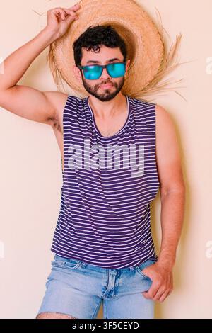 Homme élégant en été tenant un chapeau de paille et des lunettes de soleil Homme en tenue d'été, pêcheur avec un chapeau de paille. Jeune homme portant un débardeur rayé bleu Banque D'Images