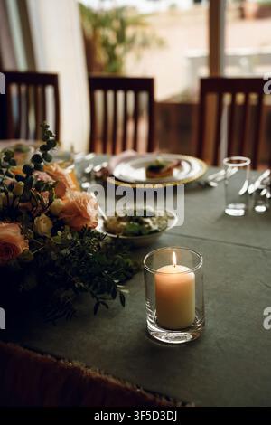 Bougie allumée dans un porte-bougie en verre sur une table dressée avec un arrangement floral, créant une atmosphère chaleureuse, confortable et romantique pour le dîner Banque D'Images