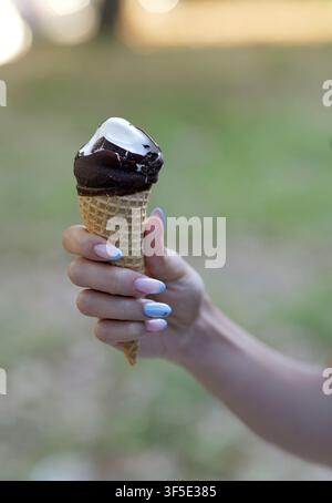 Main de femme tenant une glace au chocolat fondante, savourant un dessert froid et rafraîchissant pendant une saison chaude et ensoleillée en plein air avec de magnifiques tons pastel Banque D'Images