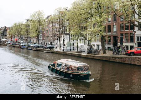 Amsterdam, pays-Bas - 18 avril 2026 : croisière touristique en bateau le long d'un canal bordé d'arbres avec des maisons hollandaises historiques dans le centre d'Amsterdam Banque D'Images