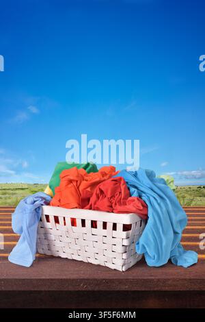 Un panier à linge en osier blanc débordant de vêtements vibrants et colorés posé sur une surface en bois devant un ciel bleu clair et un fond de paysage Banque D'Images