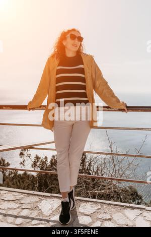 Femme au point de vue côtier, voyageuse souriante en veste jaune se tenant à une vue panoramique sur l'océan profitant du soleil éclatant du matin Banque D'Images