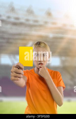 Une jeune femme indonésienne d'Asie du Sud-Est concentrée, portant un maillot orange, brandit une carte jaune comme signal d'avertissement lors d'un match sportif dans un grand, brig Banque D'Images