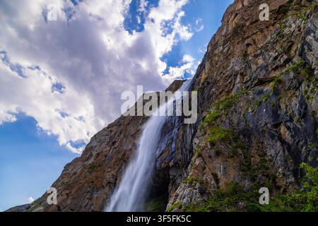 Une haute cascade dévale une montagne escarpée sous un ciel lumineux. Des nuages dramatiques accentuent la beauté naturelle de la scène sauvage. Banque D'Images