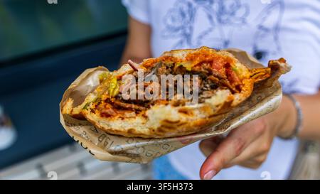 Femme tenant un sandwich shawarma croustillant à moitié mangé enveloppé dans du papier de marque en plein air, encas de rue, lumière vive, gros plan. Banque D'Images