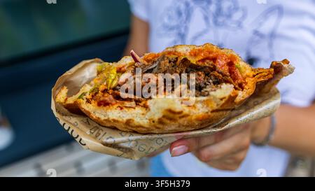 Femme tenant un sandwich shawarma croustillant à moitié mangé enveloppé dans du papier de marque en plein air, encas de rue, lumière vive, gros plan. Banque D'Images