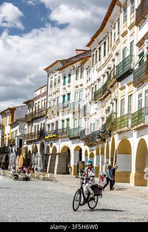 Place historique ensoleillée à Évora, Portugal. Des bâtiments blanchis à la chaux avec des volets verts bordent des rues pavées. Un cycliste passe devant des passerelles en arc Banque D'Images