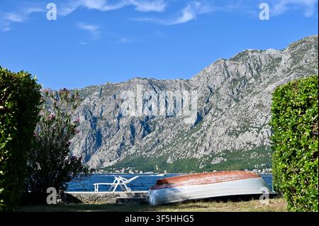 Un bateau renversé reposant sur le rivage herbeux de la côte, encadré de buissons verts avec une mer bleue époustouflante et d'immenses montagnes rocheuses sous un ciel clair. Banque D'Images