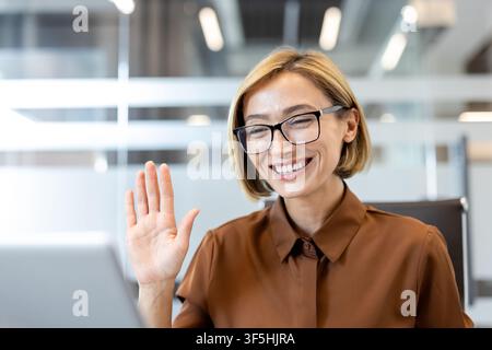 Femme d'affaires heureuse portant des lunettes et un chemisier marron, souriant et agitant la main, participant à une réunion en ligne depuis un espace de travail de bureau Banque D'Images