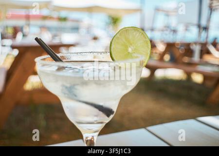 Close-up d'un cocktail Margarita rafraîchissant garni d'une tranche de citron vert fraîche et d'un bord salé, servi dans un verre classique avec des pailles noires sur une table de resort Banque D'Images
