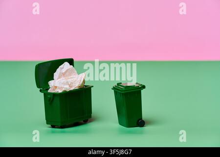Deux poubelles miniatures vertes se dressent sur une surface menthe contre un fond rose, l'une débordant de papier froissé et l'autre vide, illu Banque D'Images