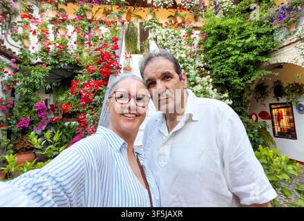 Couple mûr souriant prend un selfie dans une cour vibrante remplie de fleurs à Cordoue, en Espagne, entouré de plantes en pot colorées, d'architecture traditionnelle Banque D'Images