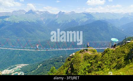 Un plan grand angle d'un long pont suspendu s'étendant sur une vaste vallée montagneuse. Plusieurs personnes portant des casques de sécurité sont vues en train de traverser Banque D'Images