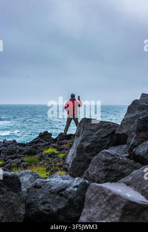 Voyageur en veste rouge vif photographiant l'océan depuis des falaises rocheuses dramatiques en Islande. Paysage côtier nuageux avec une mer turquoise Banque D'Images