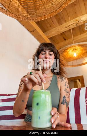 Jeune femme tatouée remue un latte matcha glacé tout en se détendant dans un café confortable aux plafonds en bois chaleureux, aux lampes suspendues tissées et aux sièges rayés. Banque D'Images