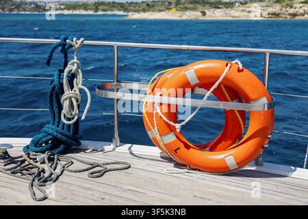 Bouée de sauvetage orange vif fixée à une rambarde en acier inoxydable sur le pont d'un yacht, avec une corde marine enroulée au premier plan et une mer bleue claire le long d'une roche Banque D'Images