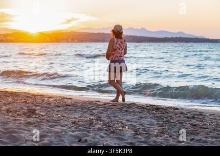 Femme en haut fleuri et chapeau marchant pieds nus le long d'une plage de sable au coucher du soleil, avec des vagues douces, une lumière dorée chaude, des montagnes lointaines et une tranquille Banque D'Images