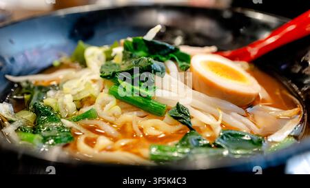 Close-up d'un bol de ramen japonais avec des nouilles dans un bouillon savoureux, garni d'un demi-œuf dur, d'oignons verts et de légumes feuillus. Plat réconfortant servi chaud dans un r Banque D'Images