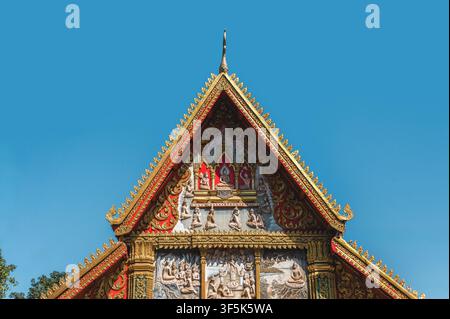 Détail de toit doré orné du temple Wat Si Muang à Vientiane, au Laos, présentant des figures bouddhistes sculptées, des pignons rouges et des pinacles décoratifs sous un cl Banque D'Images