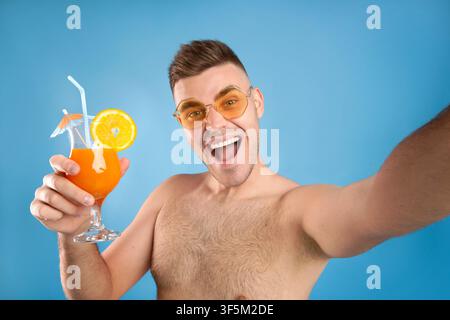 Jeune homme excité dans des lunettes de soleil tenant un cocktail d'été rafraîchissant et prise de selfie sur fond de studio bleu Banque D'Images