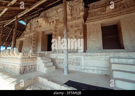 Ancien temple de pierre se dresse sous un abri en bois. Les reliefs sculptés représentent des dieux et des scènes cérémonielles. Des marches mènent à une porte obscurcie sous un mur ensoleillé Banque D'Images