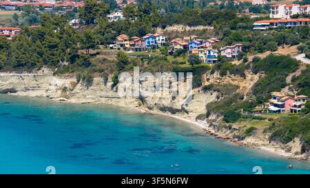 Skala Fourkas, Grèce, 11 juillet 2026. La mer turquoise rencontre des falaises rocheuses claires en dessous d'appartements et maisons résidentielles colorés entourés d'une végétation dense Banque D'Images