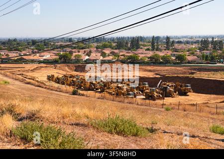 Une grande flotte de machines de terrassement est stationnée dans une excavation nivelée en dessous d'un quartier de banlieue traversé par des lignes utilitaires aériennes. Banque D'Images