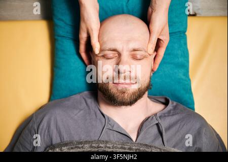 Gros plan d'un homme se faisant masser le visage thaïlandais. Masseuse faisant le traitement de massage traditionnel pour patient masculin. Client chauve avec les yeux fermés, méditant, profitant du massage des mains dans une atmosphère calme. Banque D'Images