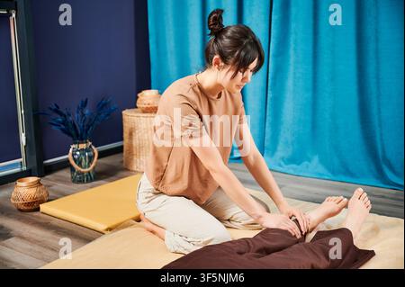 Masseuse faisant massage yoga thaïlandais. Thérapeute de femme faisant le traitement de massage traditionnel pour la patiente féminine à l'intérieur. Client allongé sur tapis, méditant, profitant d'un massage des mains dans une atmosphère calme. Banque D'Images