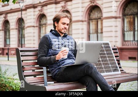 L'homme est assis sur un banc en bois dans un cadre urbain, travaillant sur un ordinateur portable avec une tasse de café à la main. Le panneau solaire repose à côté, mettant l'accent sur l'espace de travail mobile durable au milieu de l'architecture historique. Banque D'Images