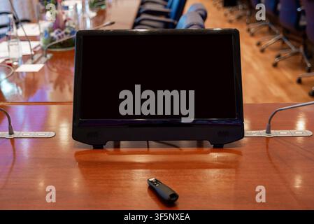 Moniteur noir posé sur une table en bois dans une salle de conférence. Une petite clé USB repose près de la base. Des chaises et des microphones suggèrent une réunion d'affaires. La scène implique Banque D'Images