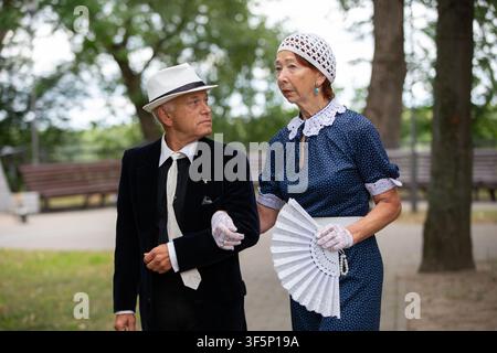 Un couple âgé marche main dans la main dans un parc, vêtu de vêtements vintage. Banque D'Images