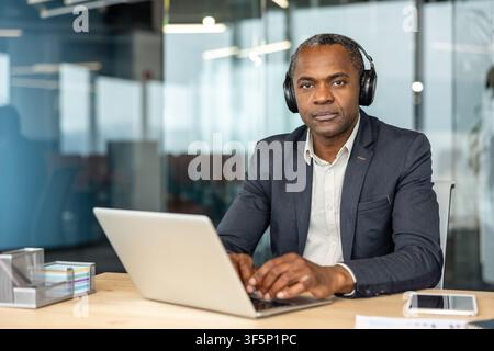 Homme professionnel portant des écouteurs et un costume sombre, assis à un bureau en bois et travaillant assidûment sur un ordinateur portable dans un environnement de bureau d'entreprise contemporain, concentré sur la productivité et la connectivité Banque D'Images