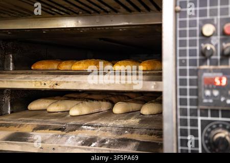 Du pain frais cuisant dans un four commercial avec des pains dorés sur les étagères supérieures et de la pâte crue posée sur les grilles inférieures tandis que le panneau de contrôle numérique Banque D'Images