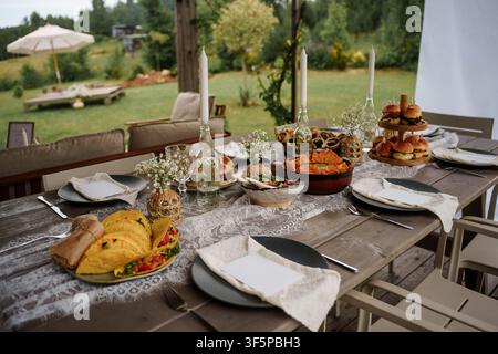 Table rustique en plein air dressée pour une fête estivale dans le jardin avec des tacos, des sliders et une décoration élégante aux bougies dans un cadre naturel de jardin Banque D'Images