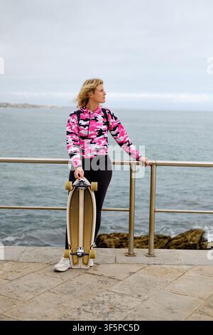 Femme en sweat à capuche camouflage rose tenant une planche à roulettes sur une promenade côtière en regardant l'océan Banque D'Images